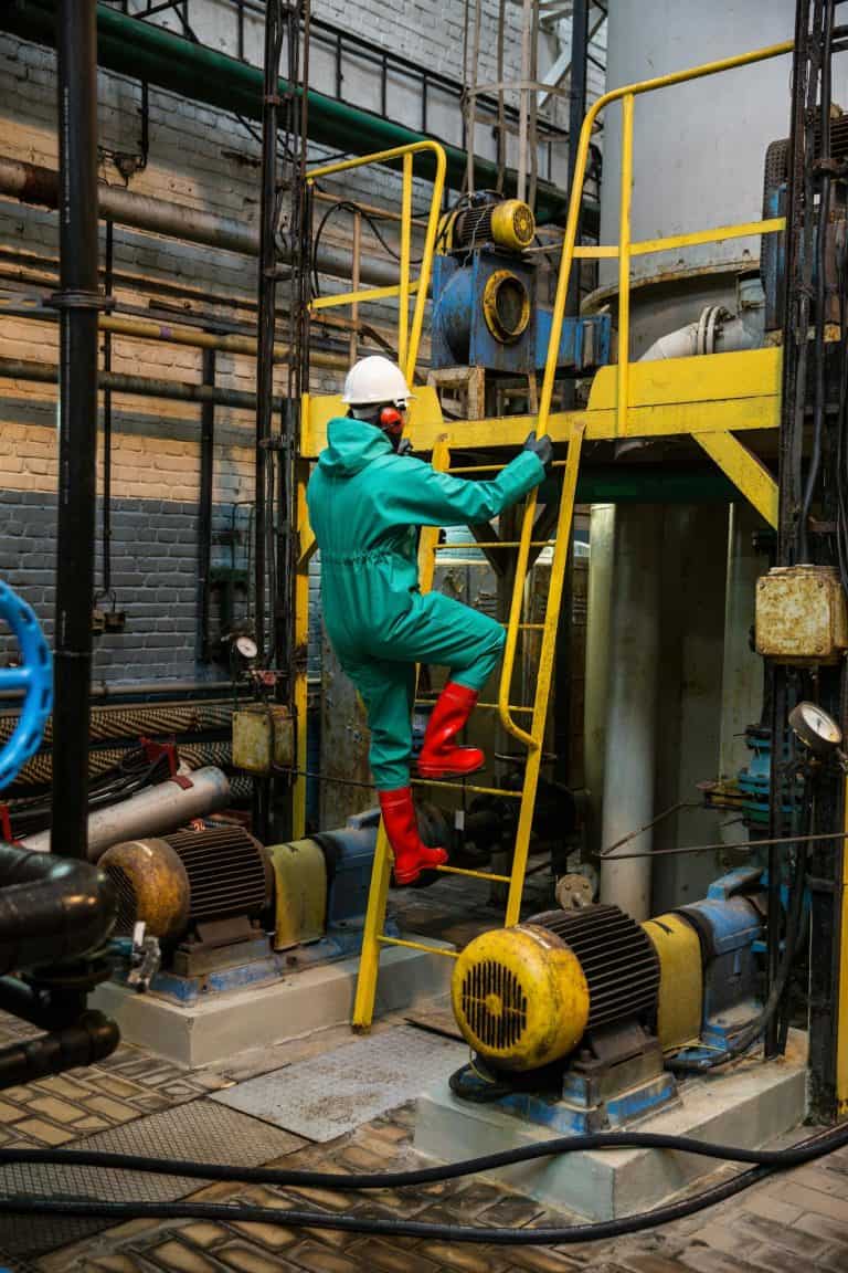 A worker in safety gear climbs a yellow ladder in an industrial factory setting, surrounded by machinery.