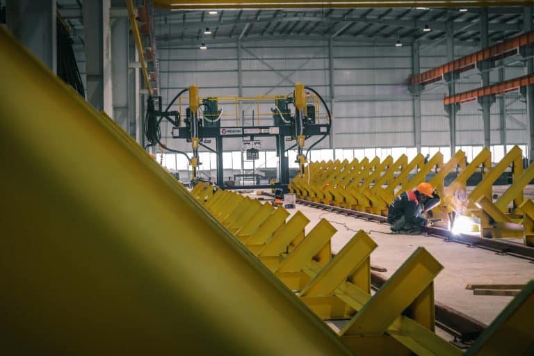 A worker welding metal in an industrial warehouse with heavy machinery.